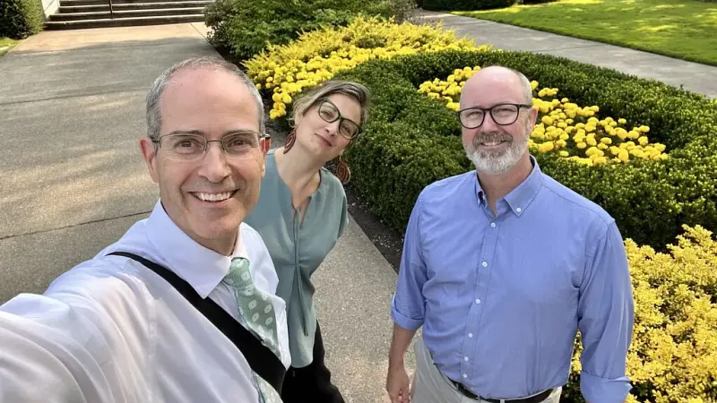 A selfie with Chris Long, Nicky Agate, and Jason Rhody in front of Johnson Hall at the University of Oregon with a hedge in the shape of an O and yellow flowers in the middle